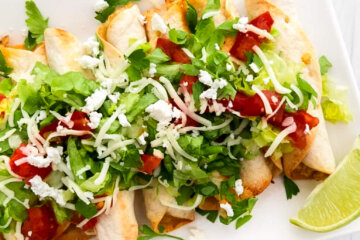 A rectangular platter of baked chicken taquitos topped with lettuce, shredded cheese and diced tomatoes and served with lime wedges.
