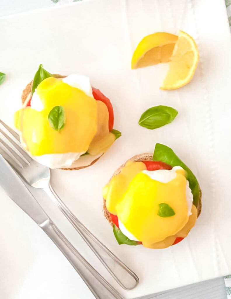 Plate of Caprese Eggs Benedict topped with fresh basil and served with lemon wedges.