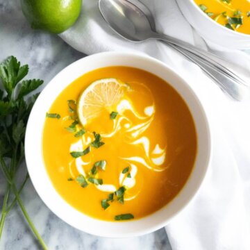 Bowl of Coconut carrot soup swirled with coconut milk and topped with fresh herbs and lime wedge.