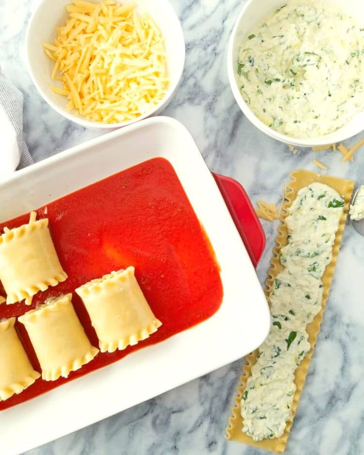 Lasagna noodle with cheese mixture spread overtop next to a baking dish with some rolled up lasagna rolls in tomato sauce.