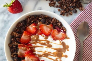 Bowl of yogurt topped with healthy dark chocolate granola and strawberries all drizzled with peanut butter.