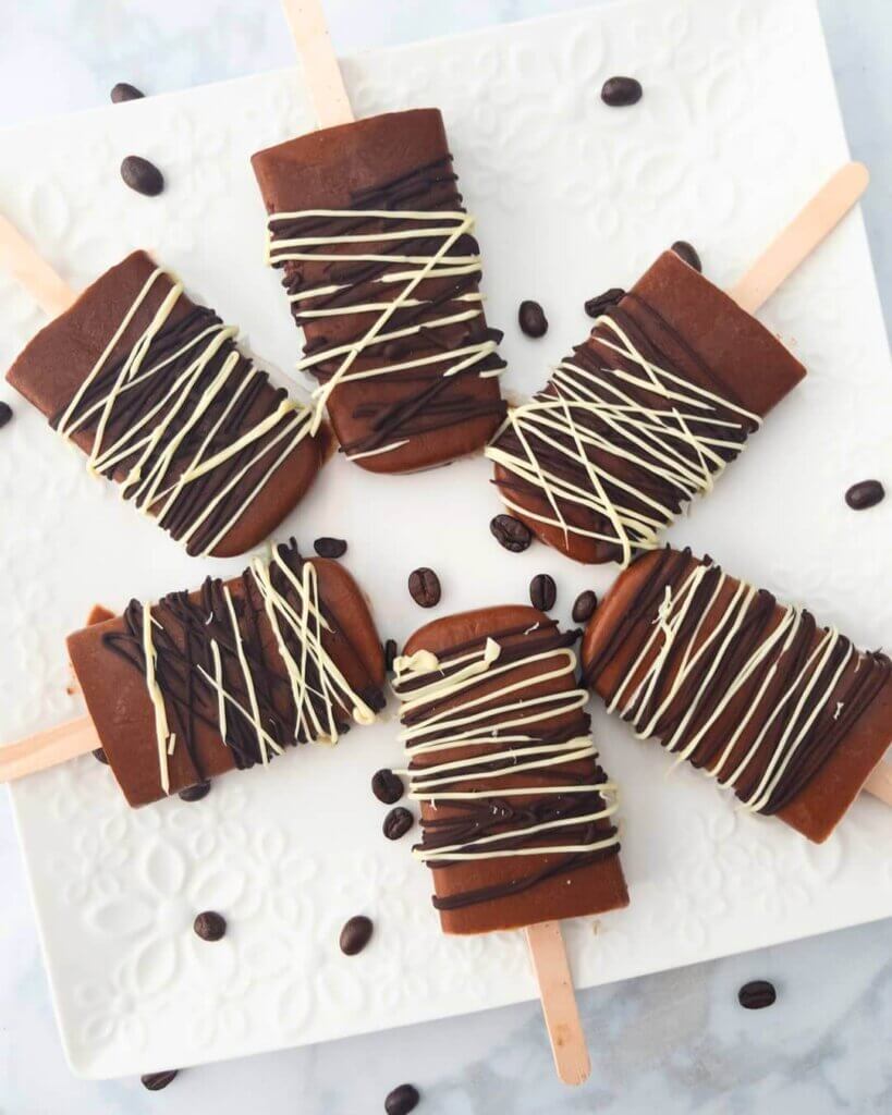 A square platter with Mocha Iced Coffee Popsicles drizzled with dark and white chocolate and surrounded by coffee beans.