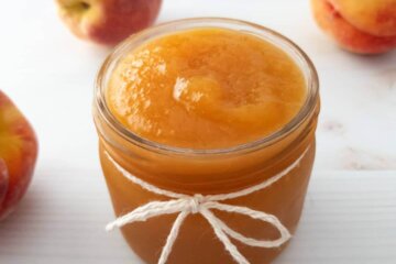A jar of Homemade Peach Jam surrounded by fresh peaches.