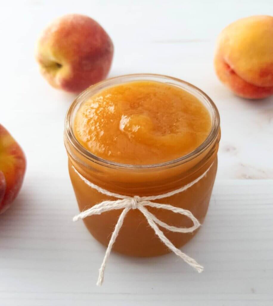A jar of Homemade Peach Jam surrounded by fresh peaches.