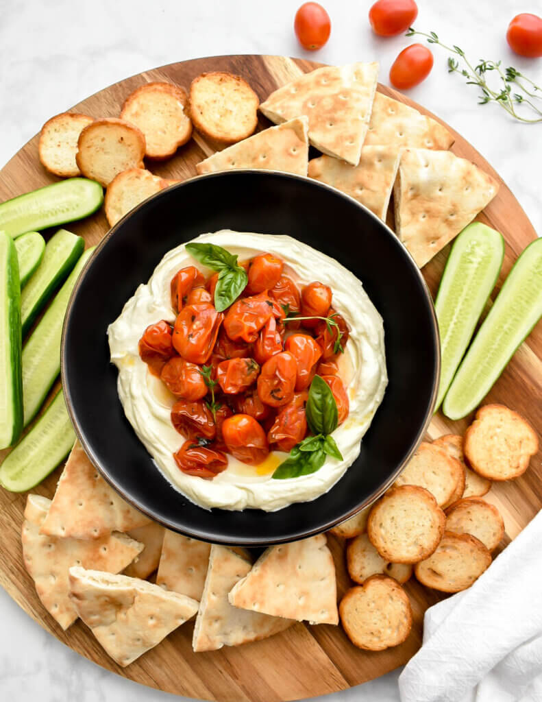 A bowl of whipped feta topped with roasted grape tomatoes served with pita, crackers and cucumbers on a wooden platter.