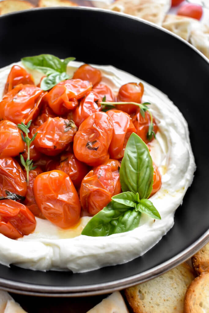A bowl of whipped feta dip topped with roasted grape tomatoes and fresh basil and thyme.
