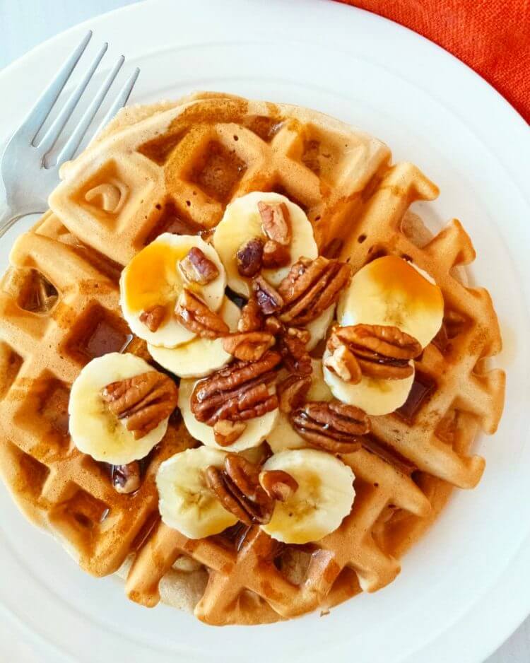 Plate with a Healthy Banana Nut Waffle topped with banana slices, pecans and drizzled with maple syrup.
