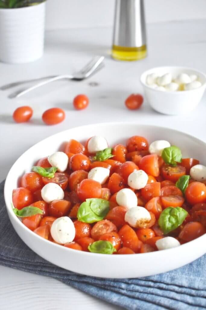 Bowl of Cherry Tomato Caprese Salad on a kitchen table with extra ingredients around it.