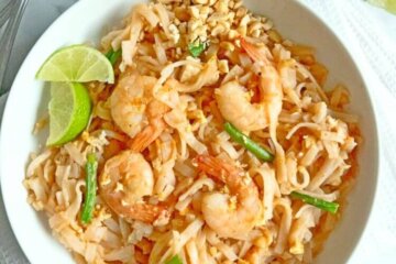 A bowl of Shrimp Pad Thai set on a white napkin and topped with lime wedges.