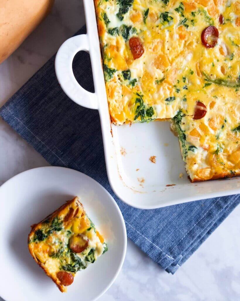 A Sausage, Squash and Spinach Breakfast Bake in a baking dish next to a plated slice.