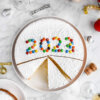 A Greek Vasilopita New Years Cake decorated with m&m's spelling out the year 2023 sliced on a platter with a slice on a small plate next to it.