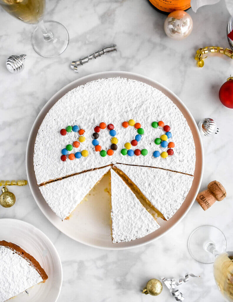 A Greek Vasilopita New Years Cake decorated with m&m's spelling out the year 2023 sliced on a platter with a slice on a small plate next to it.