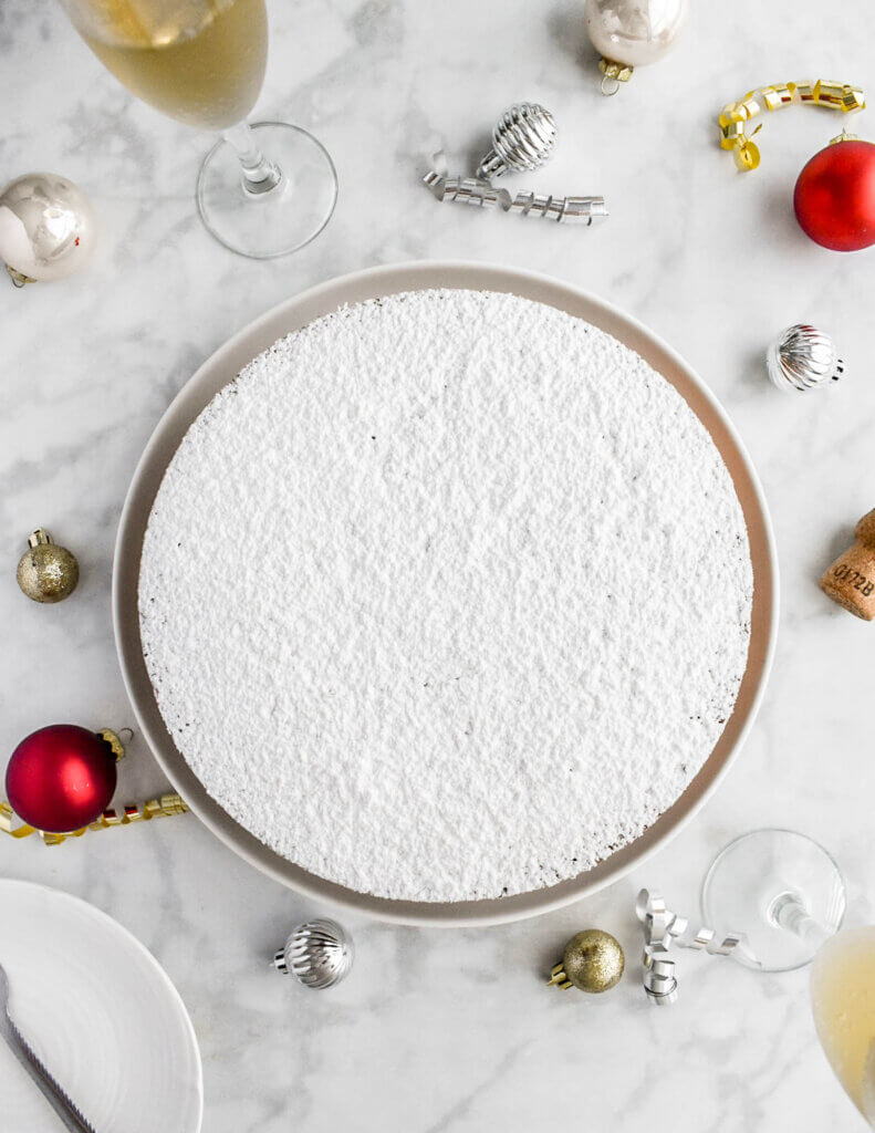 A vasilopita cake sprinkled with icing sugar and set on a grey marble countertop surrounded by glasses of champagne, ornaments and party streamers.