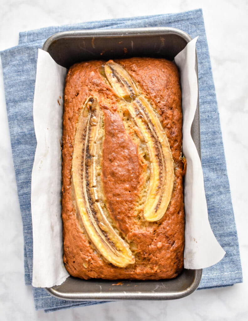 A freshly baked loaf of banana bread topped with banana slices in a loaf pan set on a blue kitchen towel.