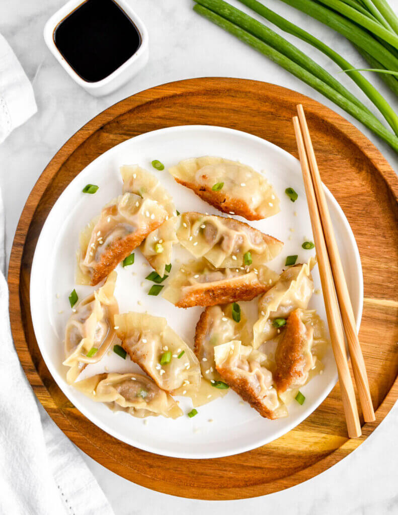 Pork pot stickers on a white plate set down next to a bowl of soy sauce and chopsticks.