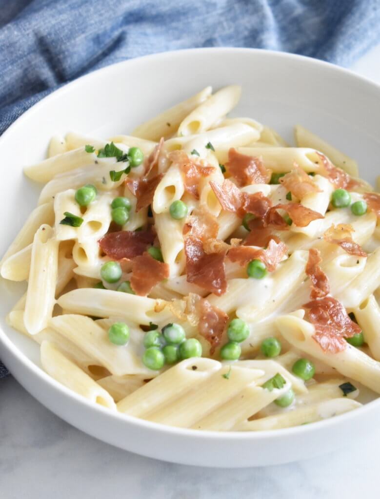 A bowl of Creamy Penne with Peas and Crispy Prosciutto on a marble counter.