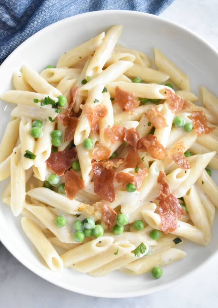 Topview of a plate of creamy pasta with crispy prosciutto and peas set on a table.