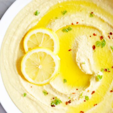 Closeup of a bowl of hummus topped with olive oil, lemon slices and red pepper flakes.