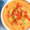 roasted red pepper hummus in a bowl with chopped red pepper garnish and fresh herbs.