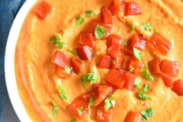 roasted red pepper hummus in a bowl with chopped red pepper garnish and fresh herbs.