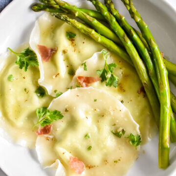 A plate of asparagus ricotta ravioli topped with crispy prosciutto and fresh herbs and served with fresh asparagus.