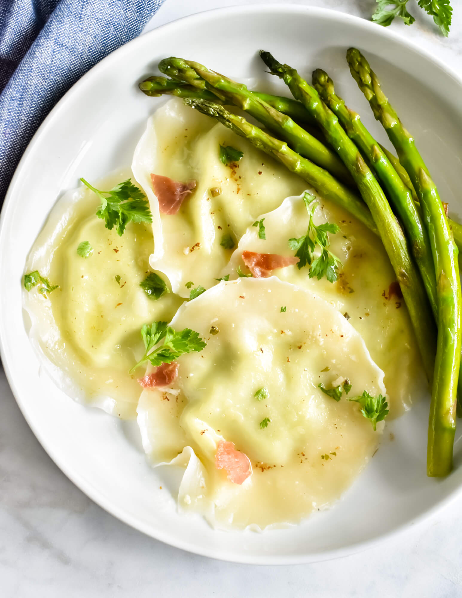 A plate of asparagus ricotta ravioli made with wonton wrappers and topped with crispy prosciutto and fresh herbs served with fresh asparagus set next to a blue cloth napkin.