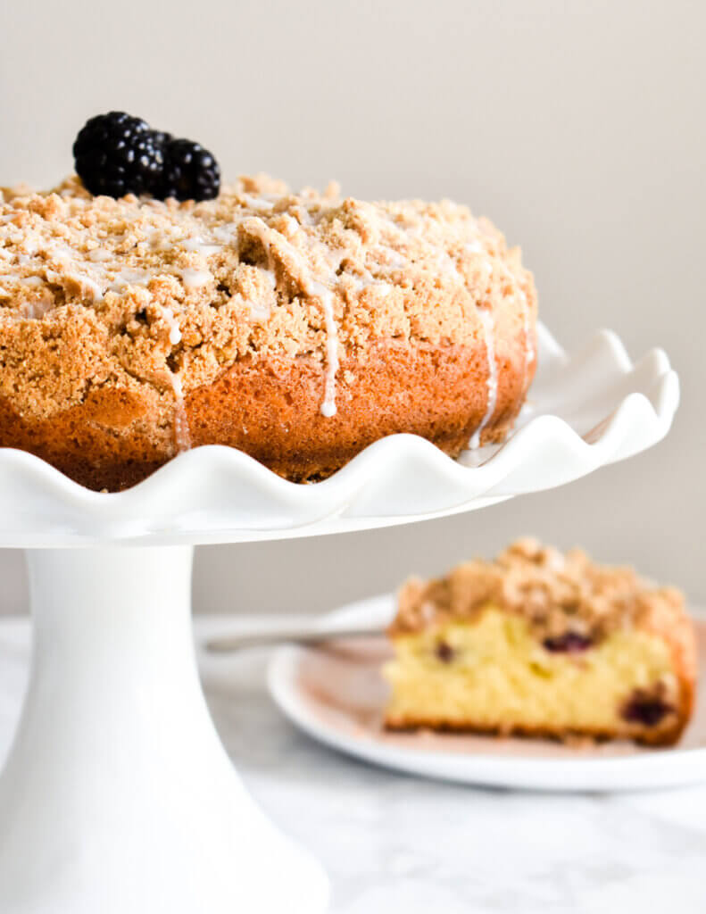 Blackberry Coffee Cake served on a white cake stand with icing dripping down the sides set next to a plate with a slice of the coffee cake.