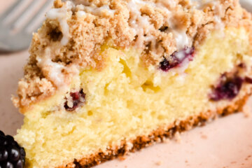 A slice of blackberry coffee cake drizzled with icing and served with fresh blackberries on a pink plate.