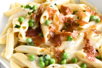 A bowl of creamy penne pasta with peas and prosciutto set next to a blue cloth napkin.