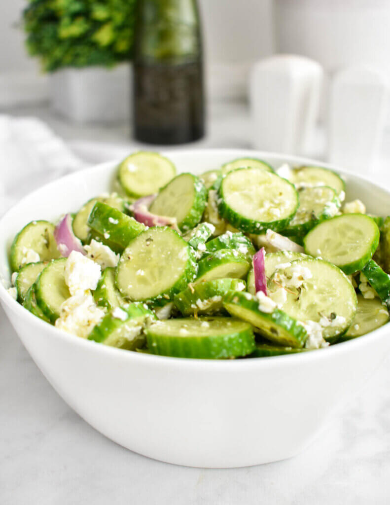 A salad made with cucumbers, feta, and red onion in a white serving bowl.