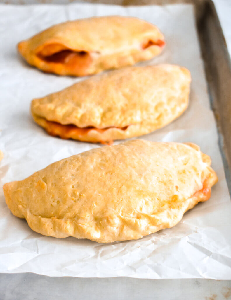 Homemade Pizza Pockets on a baking sheet lined with parchment paper that have burst to show the cheese and sauce filling oozing out.