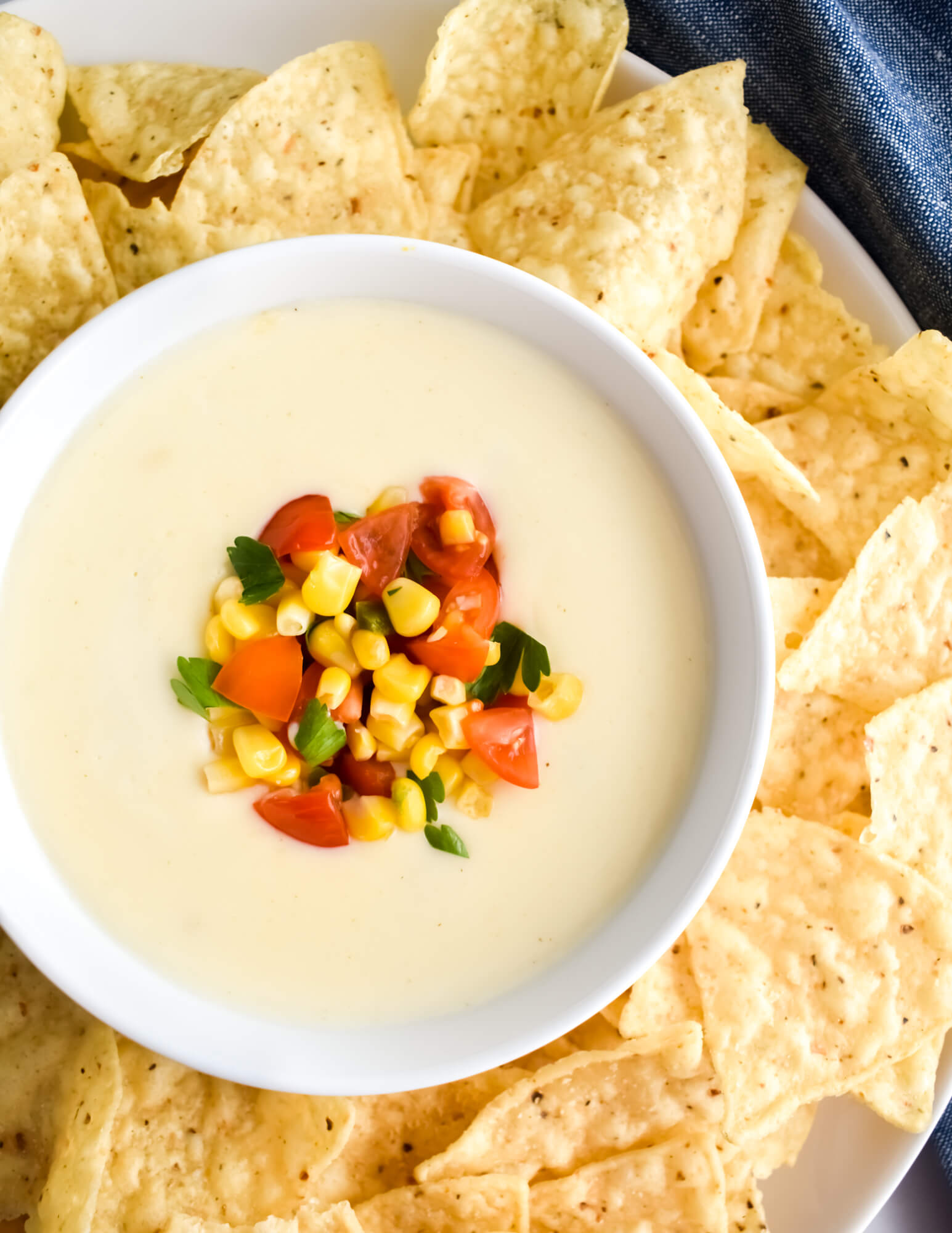 Closeup of a bowl of white queso dip topped with corn, diced tomatoes, and cilantro and nestled in a platter of tortilla chips.