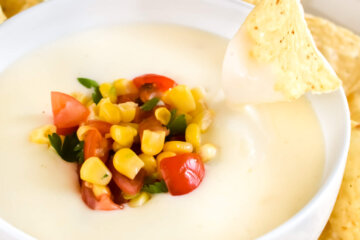 A tortilla chip being dipped into a bowl of queso blanco dip topped with fresh corn, diced tomatoes and cilantro.