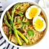 A bowl of ramen topped with grilled asparagus, eggplant and zuchini and a jammy egg.