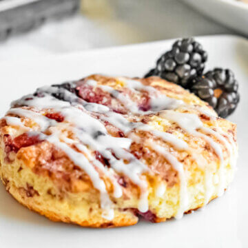 A blackberry scone drizzled with icing on a plate with fresh blackberries.