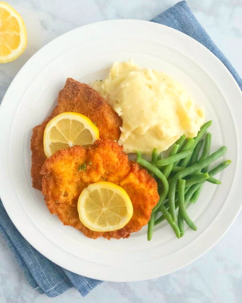 Plate of pork schnitzel topped with lemon slices and served with mashed potatoes and green beans.