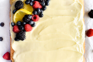 A square Triple Berry Cake topped with lemon cream cheese frosting and fresh berries and lemon slices set on parchment paper.