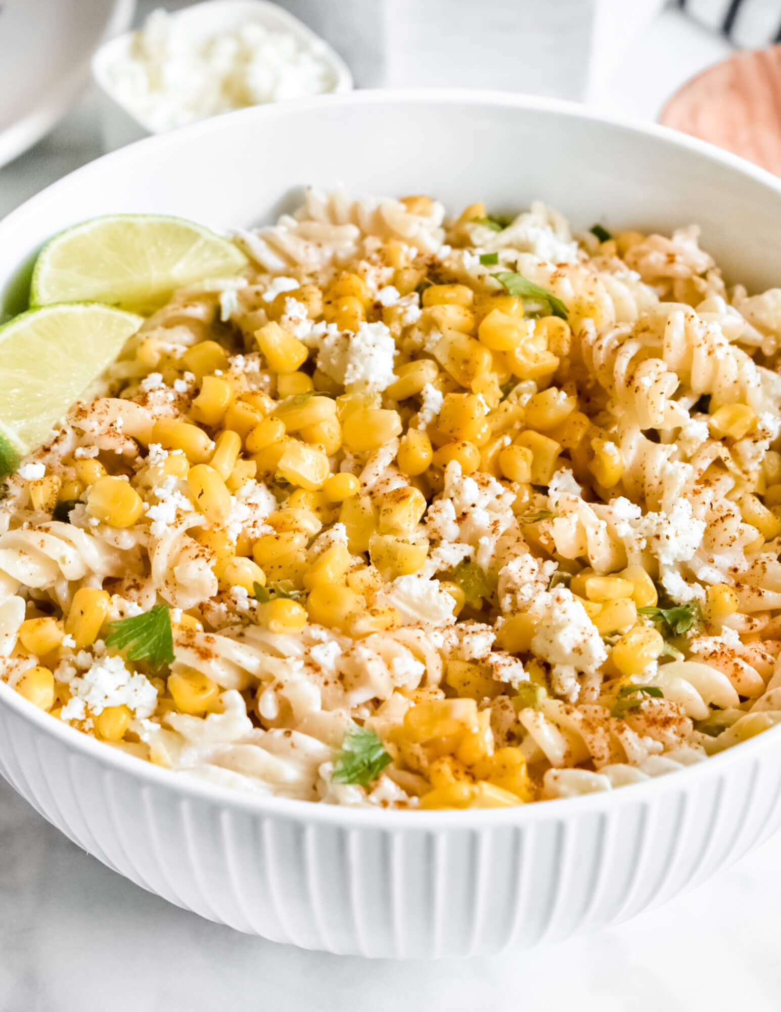 A bowl of Healthy Mexican Street Corn Pasta Salad topped with lime wedges and set on a grey marble counter in front of a bowl of cotija cheese.