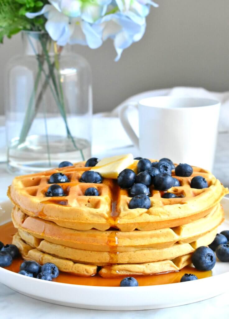 Closeup of a stack of sweet potato waffles with blueberries, maple syrup and a pat of butter on top.