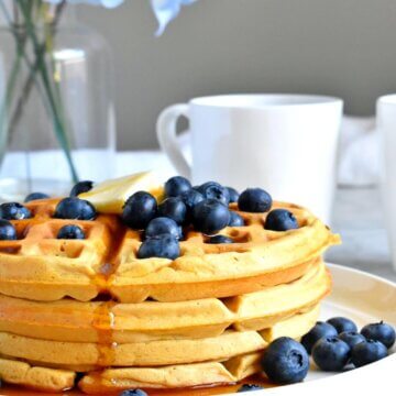 A stack of Healthy Sweet Potato Waffles topped with fresh blueberries and maple syrup with a cup of coffee.