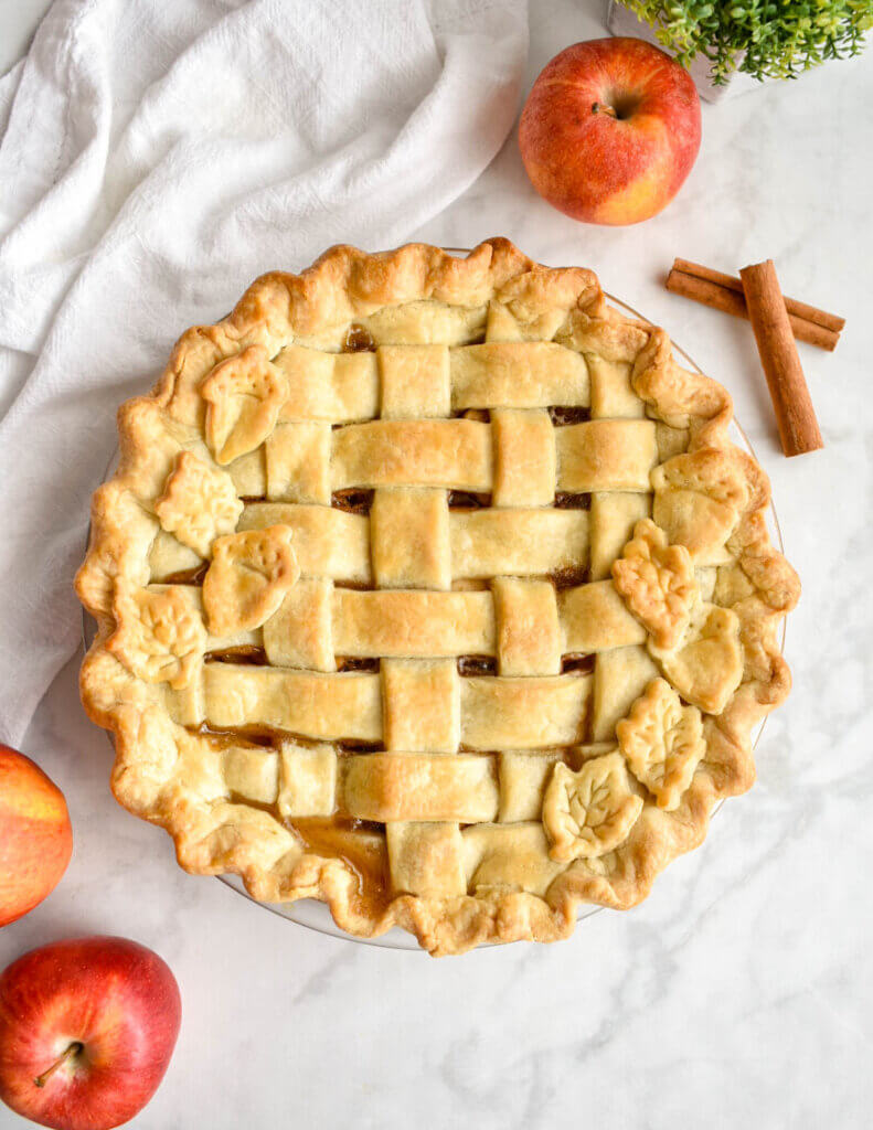 A baked apple pie with a lattice crust set on a marble countertop with apples and cinnamon sticks.