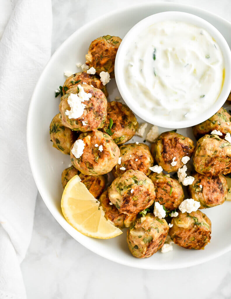 Closeup of a platter of greek chicken meatballs topped with feta cheese and served with lemon wedges and tzatziki.