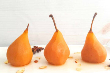 3 poached pears standing on a rectangular plate sprinkled with almond slivers.