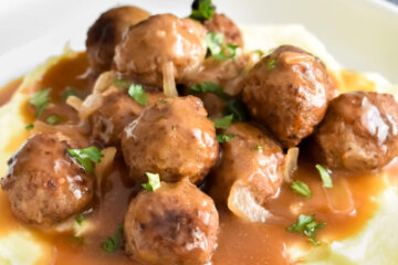 Salisbury steak meatballs piled onto whipped potatoes and topped with brown onion gravy and parsley set on a white plate next to a blue cloth napkin.