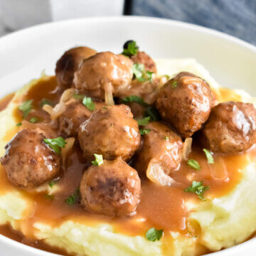 Salisbury steak meatballs piled onto whipped potatoes and topped with brown onion gravy and parsley set on a white plate next to a blue cloth napkin.