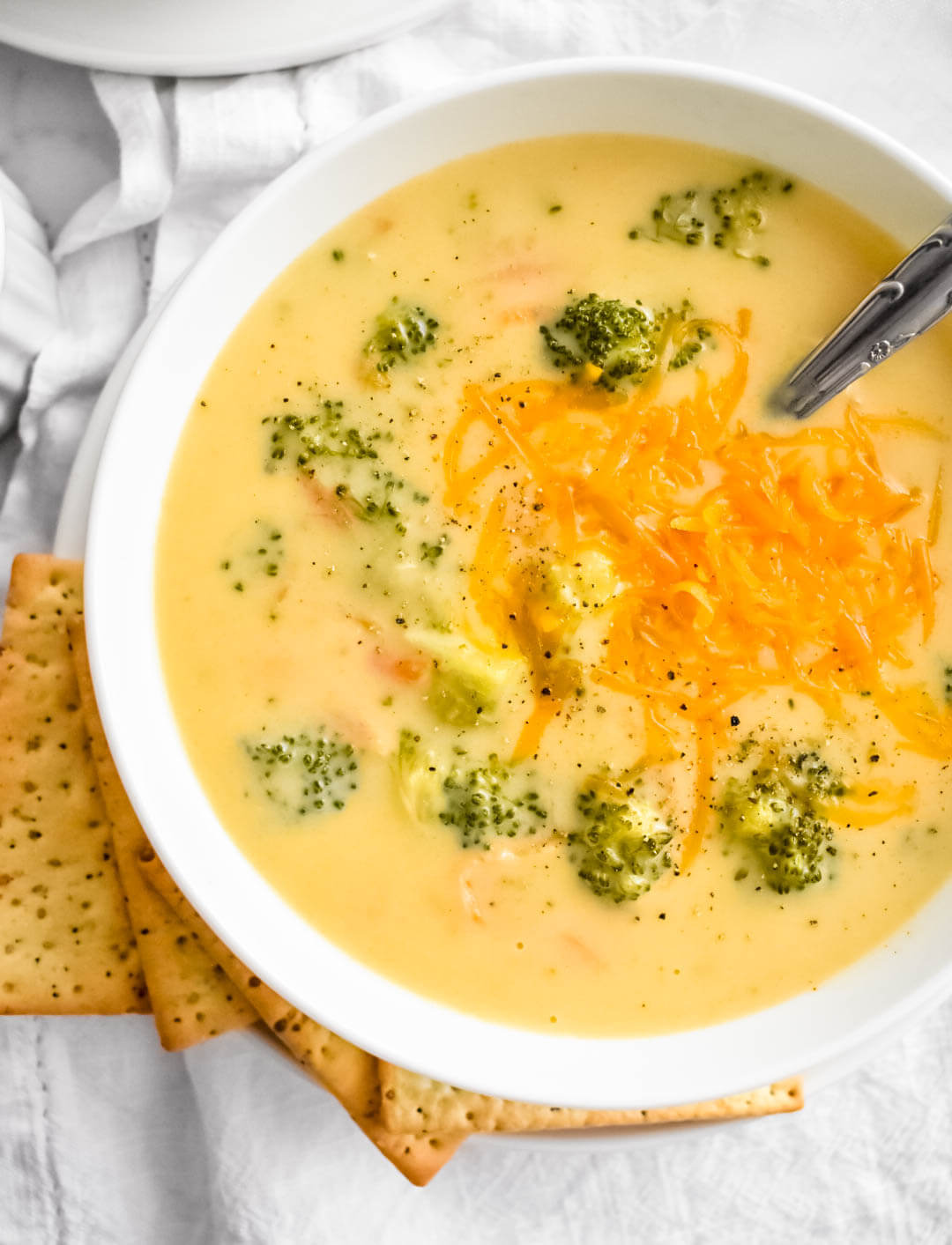 Closeup of a bowl of broccoli cheese soup topped with finely shredded cheddar and set on a white napkin next to crackers.