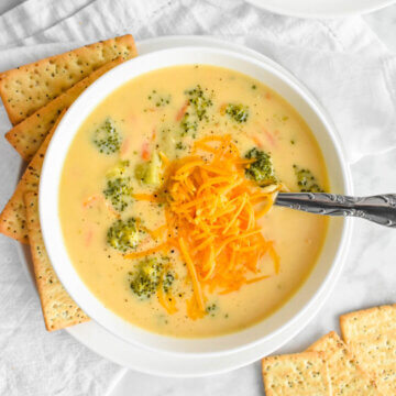 A bowl of Broccoli Cheddar Soup topped with shredded cheddar cheese and set on a white linen napkin surrounded by crackers and a small bowl of shredded cheddar cheese.