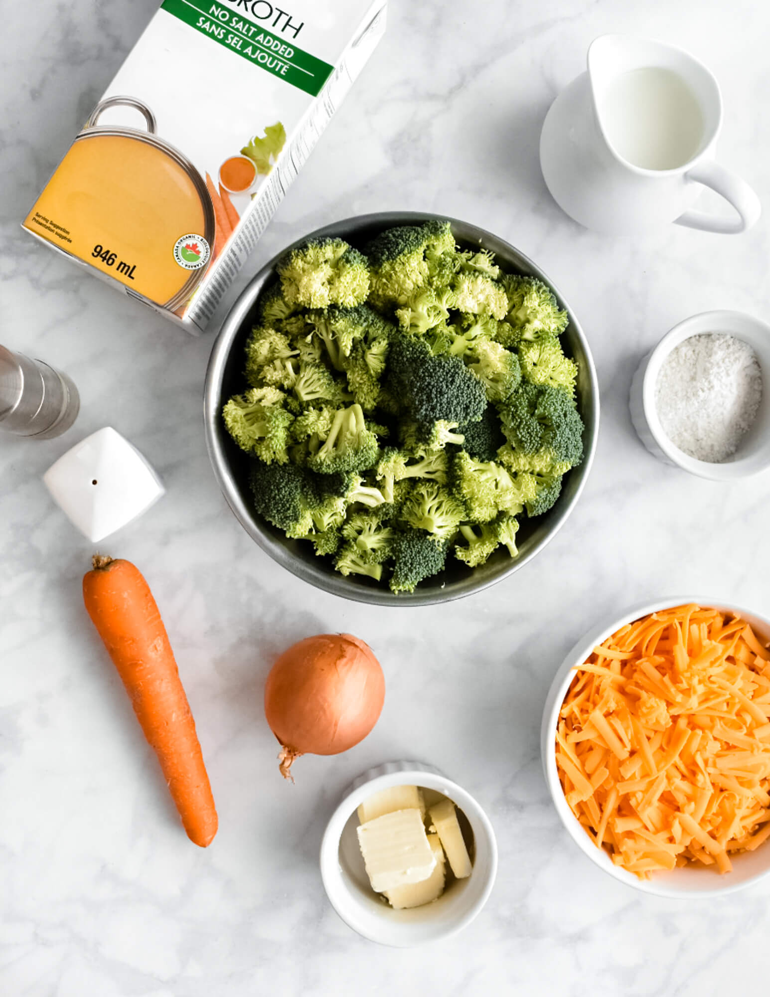 The ingredients for broccoli cheddar soup including a bowl of broccoli florets, a carrot, an onion, a bowl of shredded cheddar, butter, a bowl of flour, a small jug of milk, and salt and pepper shakers on a grey marble counter.