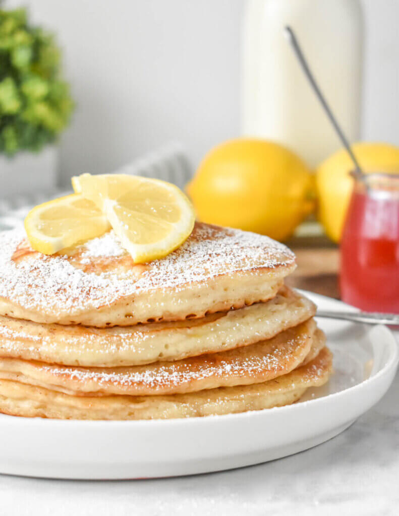 Lemon Ricotta Pancakes powdered with sugar next to a jar of strawberry syrup.
