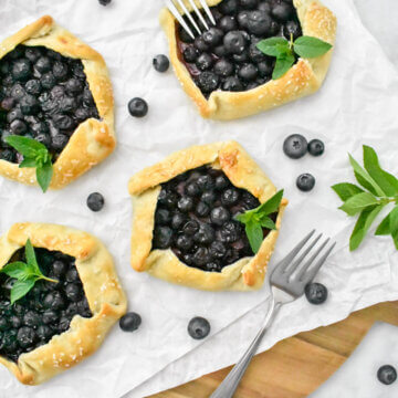 Mini Blueberry Galettes on parchment paper surrounded by fresh blueberries, mint and forks.